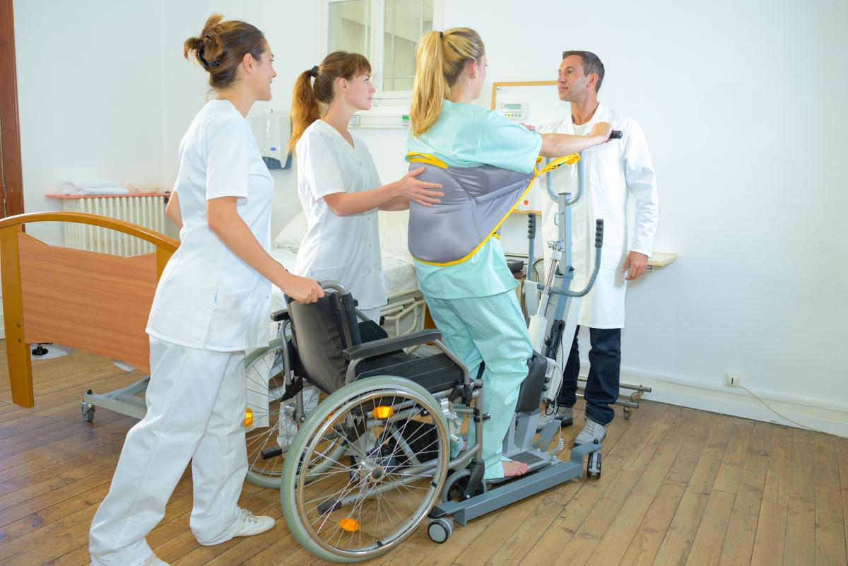A standing hoist and sling operated by two support workers and clinician to sit to stand from a manual wheelchair