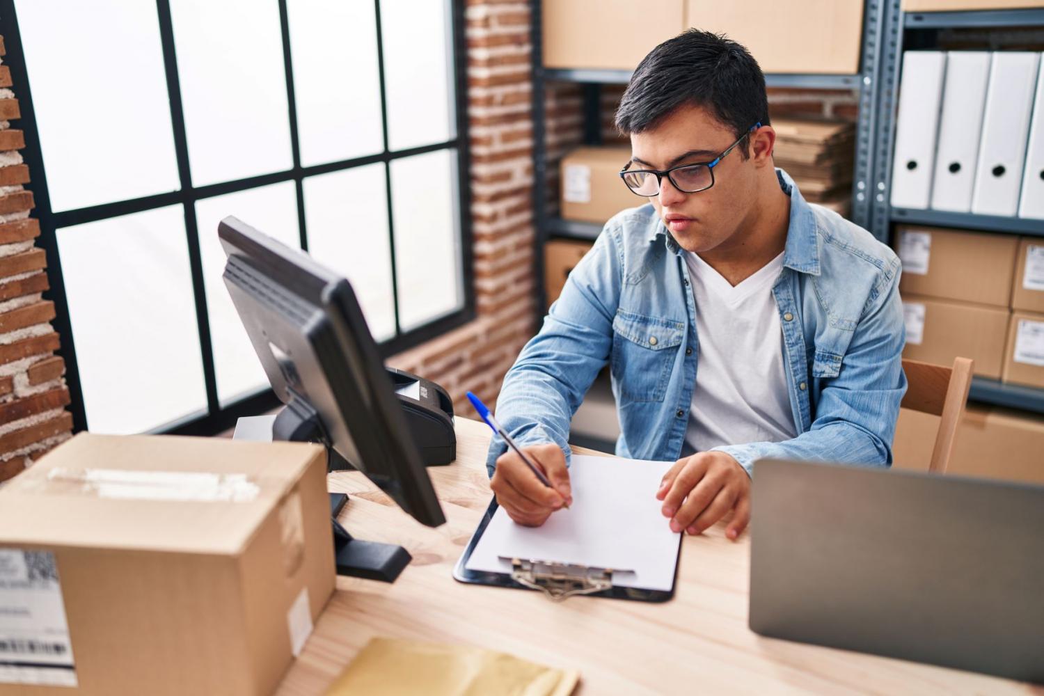 Man seated at desk writing notes on clipboard featuring computer and laptop