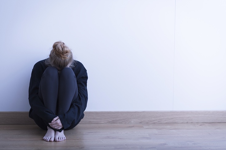a woman sits on the floor against a white wall in the corner of a photo with her head on her knees and arms clutching her legs