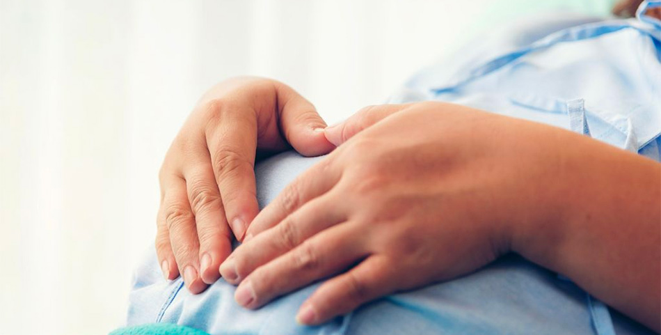 A pregnant woman wearing a light blue shirt rests her hands on her belly. We cannot see her face.