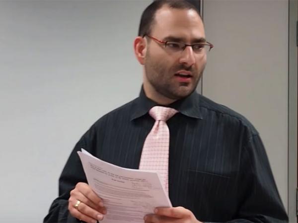 Paul standing speaking holding his notes and wearing a pink tie